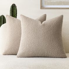 two beige pillows sitting on top of a white couch next to a green cactus plant