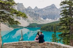 a man and woman are sitting on a stone wall looking at the lake in the mountains