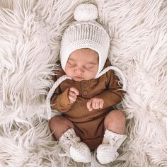 a baby is laying down wearing a brown outfit and a white knitted hat with two pom - poms