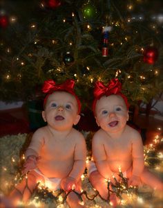 two babies sitting next to each other in front of a christmas tree with lights on it