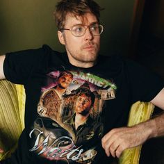 a man with glasses sitting on a couch wearing a t - shirt that has the image of two men in front of him