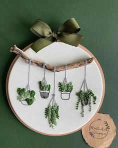 a cross stitch pattern with potted plants hanging on a wooden frame and a green ribbon