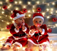 two babies dressed as santa clause sitting next to each other on a bed with christmas lights