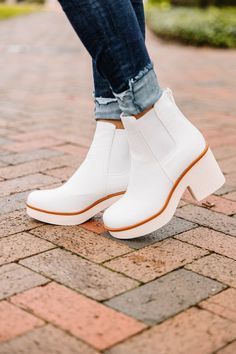 a close up of a person wearing white boots and jeans on a brick walkway with trees in the background