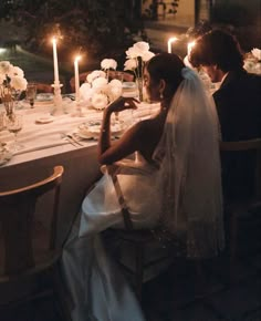 a bride and groom sitting at a dinner table with candles in the dark behind them