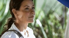 a woman in white shirt and brown bag looking off into the distance with palm trees behind her