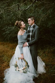 a man and woman standing next to each other in the grass with trees behind them