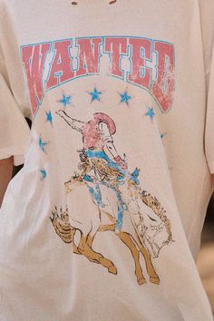 -Color: Ivory -Distressed washed graphic t-shirt -Vintage-style cowboy bull-riding and stars graphic print with "Wanted" text print -Round neckline -Short sleeves -Dropped shoulders -Torn and distressed detailing at neckline, cuffs, and hem -Oversized fit -Content: 100% Cotton -Made in Guatemala -Model is 5'9" and wearing a size Small Stars Graphic, Western Designs, Pink Cowboy, Riding Shirts, Bull Riding, Text Print, Western Design, Floral Tee, Color Ivory