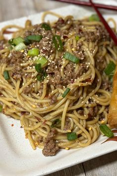 a white plate topped with spaghetti and meat next to chopsticks on a wooden table