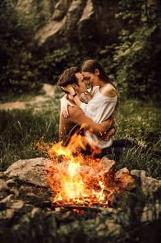 a man and woman sitting in front of a campfire with their arms around each other