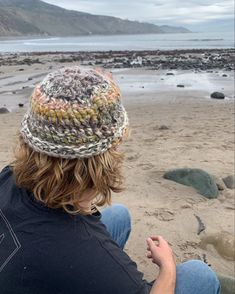 a man sitting on top of a sandy beach next to the ocean wearing a knitted hat