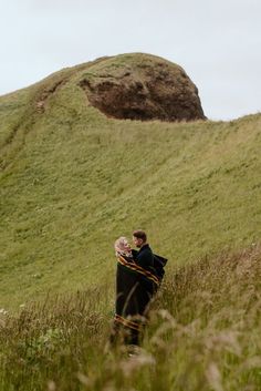 a man and woman standing in tall grass on top of a hill next to a large rock