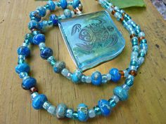 a necklace with blue and green beads on a wooden table next to a glass pendant