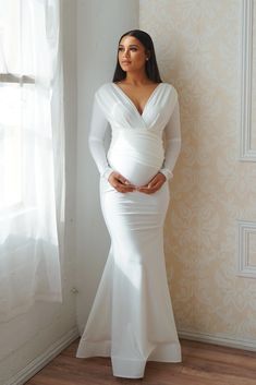 a pregnant woman wearing a white gown standing in front of a window