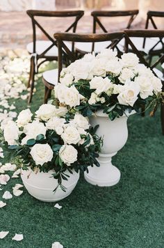 two white vases filled with flowers sitting on top of a grass covered field next to chairs