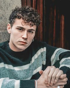 a young man with curly hair wearing a striped sweater looking at the camera while leaning against a wall