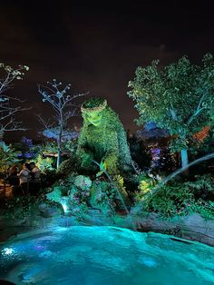 an animal statue is lit up at night in the garden with blue water and trees