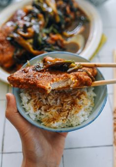 someone holding chopsticks over a bowl of rice with meat and vegetables in it