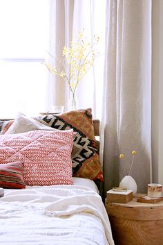 a bed sitting next to a window with white curtains and pillows on top of it