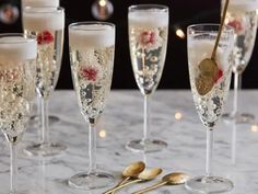 five champagne flutes are lined up on a table with gold spoons and candles in the background