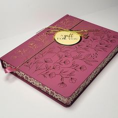 a pink book with gold foil on the front cover and floral design, sitting on a white surface