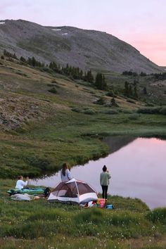 two people standing next to a tent near a body of water at sunset with mountains in the background