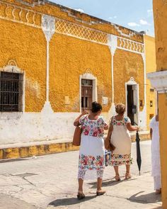 two women walking down the street in front of an old building with yellow painted walls