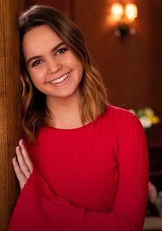 a smiling woman in a red top leaning against a wooden wall with her hands on her hips