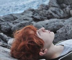 a woman with red hair laying on rocks near the ocean