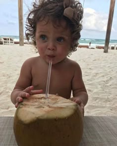 a baby sitting on the beach with a straw in it's mouth and drinking from a coconut