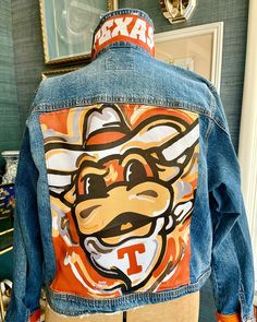 Longhorns !  A Justin Patten design has been added to a brand new medium size denim jean jacket  to create a one of a kind spirit jacket.  The collar, cuffs, and back are adorned with longhorns material.  A unique and adorable one of a kind piece.  Size medium is available for shipping tomorrow .  Pre- order available for other team jackets in any size with up to 4 weeks creation time. SilkWarrior specializes in vintage jackets upcycled with designer scarves and fabric, creating unique and styli College Jean Jacket, Painted Jacket Ideas, Custom Jean Jacket Ideas, Football Jean Jacket, Upcycle Denim Jacket, Spirit Jacket, Patten Design, Custom Jean Jacket