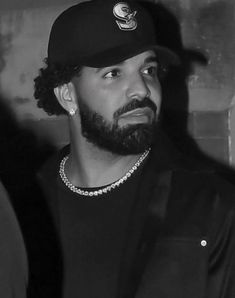 a black and white photo of a man with a beard wearing a baseball cap, standing in front of a brick wall