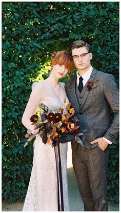 a man and woman standing next to each other in front of a hedge covered wall