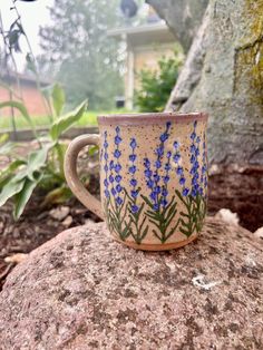 a coffee cup with blue flowers painted on it sitting on a rock in the woods