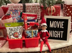 an elf is sitting in front of christmas presents on the table next to a sign that says movie night