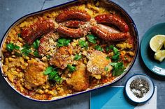 a blue dish filled with sausages and rice next to lemon wedges on a table