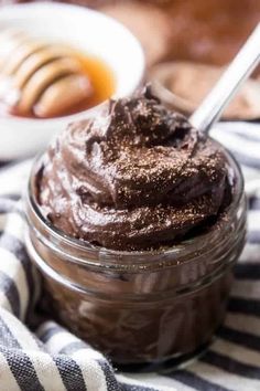 a glass jar filled with chocolate frosting on top of a striped table cloth next to a bowl of cookies