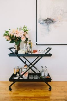 a bar cart with drinks and flowers on it in front of a wall mounted painting