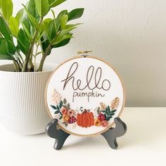 a potted plant sitting next to a cross stitch hoop with the word hello pumpkin on it