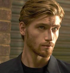 a close up of a person wearing a suit and tie with long hair in front of a brick wall