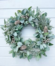 a green wreath with pine cones and greenery hanging on a white painted wall in front of a house