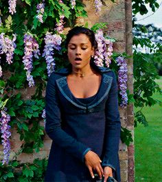 a woman standing in front of a brick wall with purple flowers growing on it's side