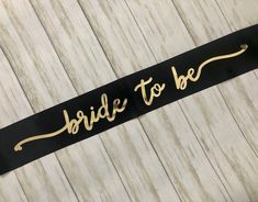 the bride to be sash has gold lettering on it and is hanging from a wooden wall