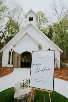 a sign sitting in front of a white church with flowers on it's easel