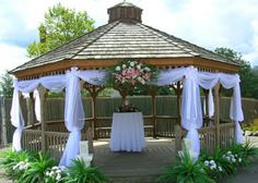 a gazebo with white drapes and flowers on the table in front of it