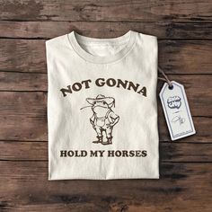 Elevate your wardrobe with a touch of humor and style with our Not Gonna Hold My Horses Shirt, Western Shirt, Cowgirl Shirt, Country Music Shirt, Concert Tee, Horse Lover, Cowboy, Silly Shirt, Funny Crafted for both comfort and sustainability, this medium-weight tee (5.3 oz/yd² or 180 g/m²) is made from 100% high-quality cotton, offering the perfect blend of year-round wearability and durability. Designed in a classic fit, it provides a relaxed, comfortable feel that's perfect for any occasion-- Novelty Cotton Shirt With Slogan, Novelty Cotton Shirt With Screen Print, Novelty Cotton Tops With Screen Print, Novelty Cotton Shirt Pre-shrunk, Cotton Band Merch Shirt With Funny Print, Cotton Shirt With Funny Print Band Merch, Funny Country Shirts, Silly Shirt, Country Music Shirt