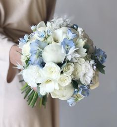 a bouquet of white and blue flowers is held by a woman's arm in a beige dress