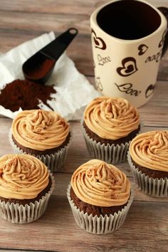 four cupcakes with frosting sitting on a table next to a coffee mug