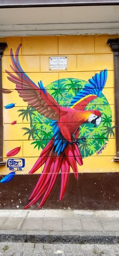 a colorful bird painted on the side of a yellow building with palm trees and leaves