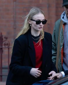 a woman in black jacket and sunglasses walking next to a man wearing a red shirt